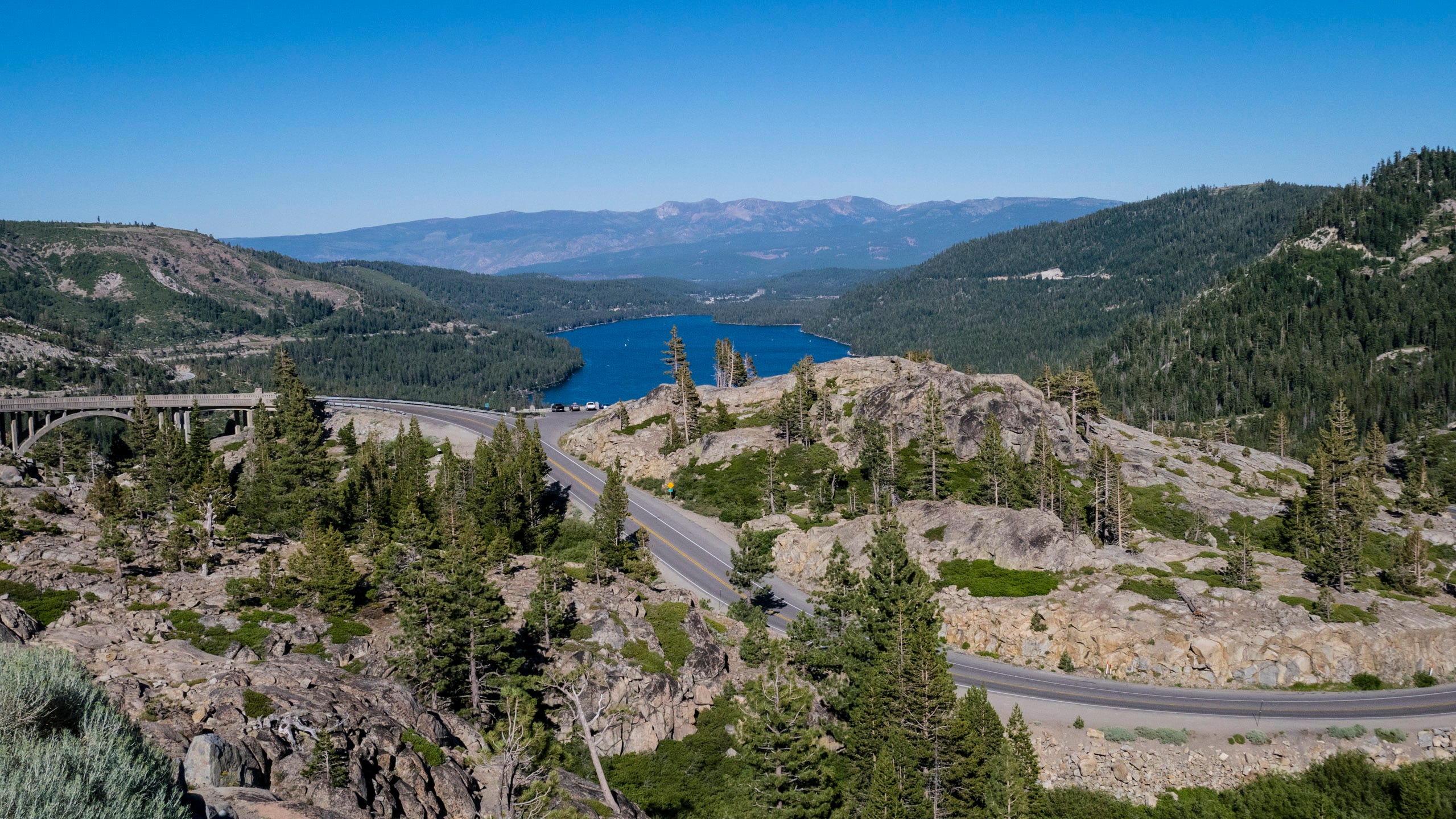 Panoramic photo looking east from Donner Pass towards a road that snakes downward among trees and rugged rock. Beyond is the deep blue of Donner Lake, surrounded by thick forests. Beyond that, in the hazy distance are more forests and the naked distant peaks of the Sierra Nevada Mountains. On the right, cutting through the forest, the grade of the Transcontinental Railroad can be seen.