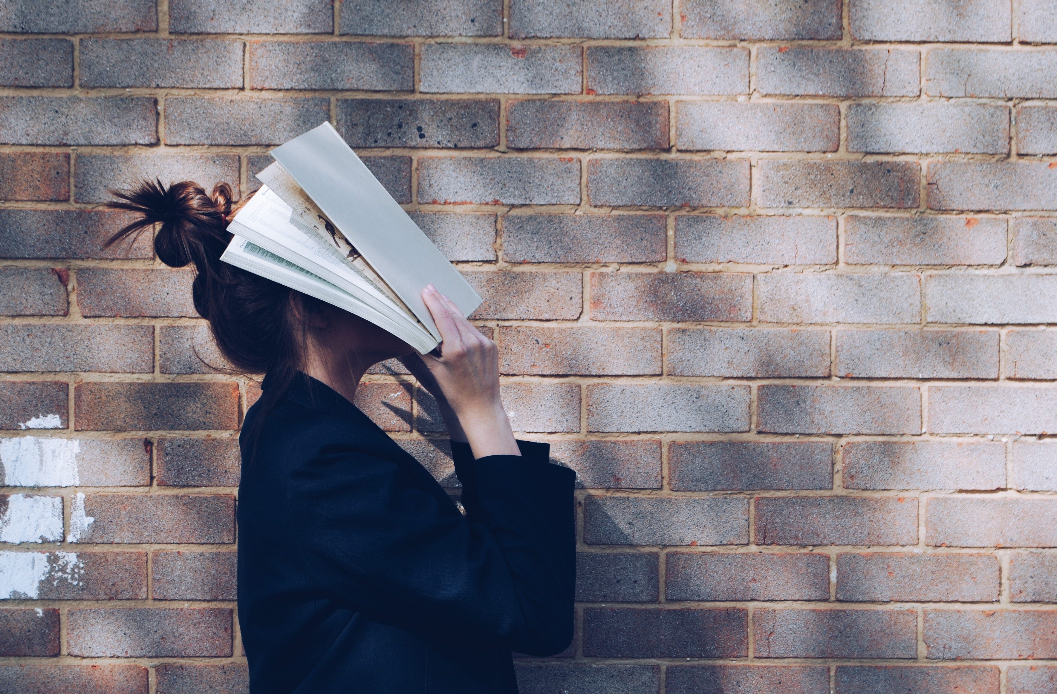 A woman stands before a brick wall. Her hair is in a bun and she is holding a book across her face.