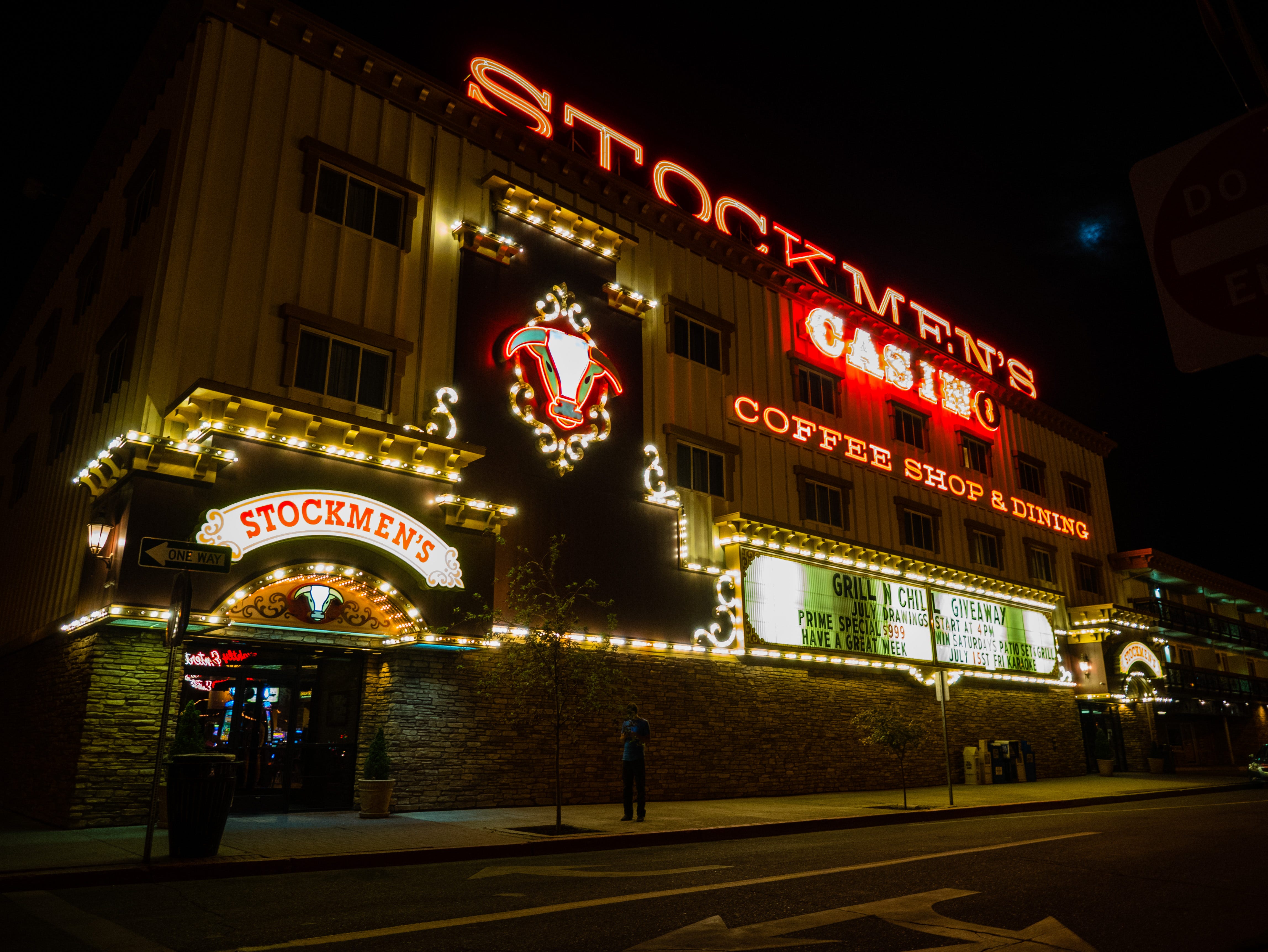 Evening photograph of the facade of the "STOCKMEN'S CASINO". The building is three plus stories tall. On top, "STOCKMEN'S" is written in oversized neon nearly the width of the entire building. Below, in letters nearly as large, is "CASINO" The letters are lit sequentially until the whole word is visible; as the photo was taken, the letters "C-A-S-I-N" are lit and the "O" is not lit. Below that, are "COFFEE SHOP & DINING" also in neon. To the left, additional lights decorate a lit head of a mult-colored steer's head looking out from the building. An additional row of lights outline the border between the ground floor and the floors above. And on the corner of the building, at the entrance, is a curved, lit, banner, "STOCKMEN'S". A giant marque has the words, "GRILL N CHILL GIVEAWAY / JULY DRAWINGS START AT 4PM / PRIME SPECIAL $9.99 WIN SATURDAYS PATIO SET AND GRILL / HAVE A GREAT WEEK / JULY 15ST FRIDAY KARAOKE". In front of the building, a man stands on the sidewalk, appearing to look at his phone.