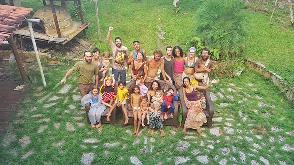 Foto da comunidade aldeia, criada por Gabriel Dread. 12 adultos e 14 crianças de variadas idades, felizes, em um jardim todo verde.