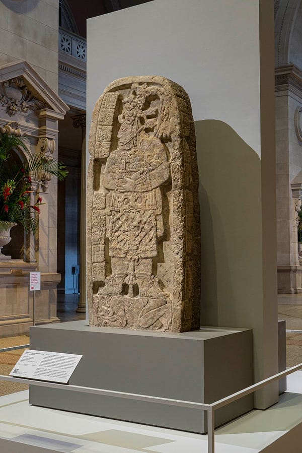 A rounded Mayan stone stela carved to represent an ancient queen. She stands in profile atop a captive who crouches below her feet. The stela stands inside The Met's Great Hall.