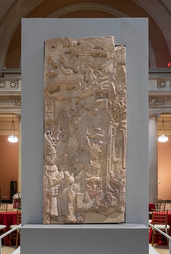 A rectangular Mayan stone stela carved to represent an ancient king. He rises to meet a visitor who enters from the left of the frame. The stela stands inside The Met's Great Hall.