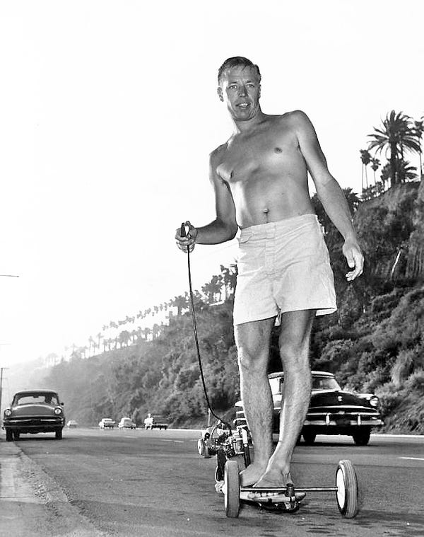 Hobie Alter rides his skateboard alongside the road. Note: Alter has added a small electric motor onto his skateboard and controls the board with a hand-held switch. Photo dated: May 11, 1965.