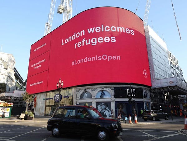 Piccadilly Lights billboard with message saying "London welcomes refugees #LondonIsOpen". Black cab is driving past beneath. 