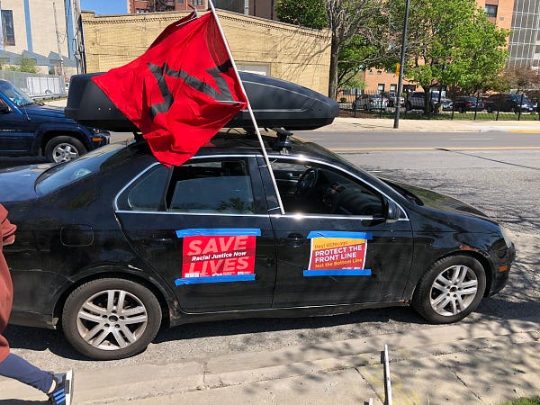 A black car from the NNU car caravan with a red and black NNU flag and NNU placard taped to the side "racial justice saves lives" and "Hey Uchicago, protect the front line, not the bottom line"
