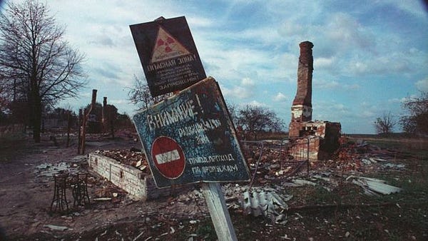 Foto de uma paisagem devastada na cidade de Chernobyl. Árvores sem folhas e construções caindo aos pedaços podem ser vistas ao fundo. No centro da imagem há uma placa enferrujada com uma frase em russo e o símbolo de "alerta de radiação". 