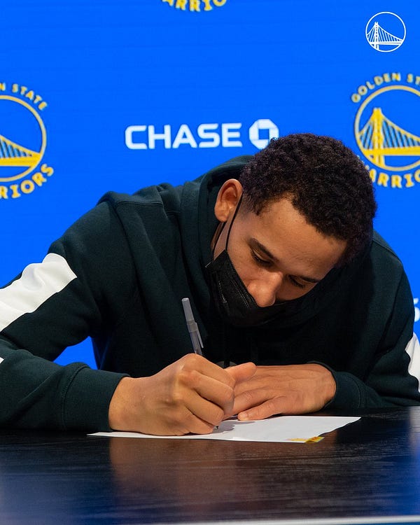 Juan Toscano-Anderson signs his contract with the Golden State Warriors in front of a Warriors backdrop.