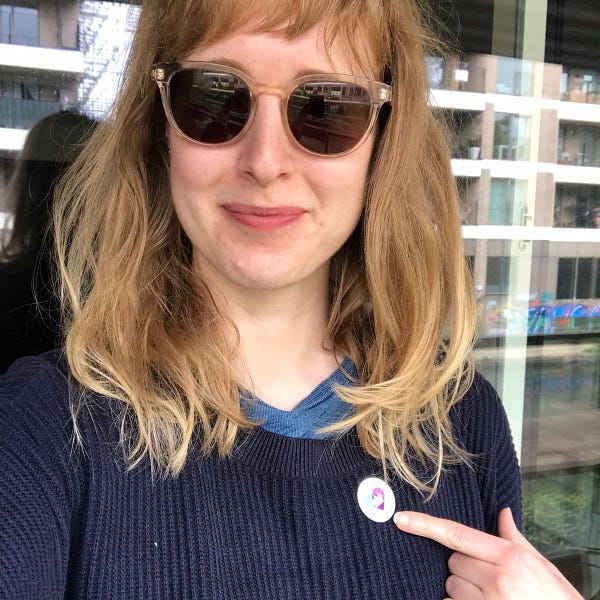 Selfie of Emily, a white blonde woman wearing sunglasses, pointing at her vaccination sticker