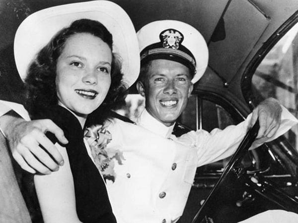 Young Jimmy Carter wearing his Naval dress uniform and hat sitting behind the wheel of a car, one arm around his young bride, Rosalynn Smith Carter. Rosalynn wears a white and navy dress with a white wide-brimmed hat. Both are smiling.