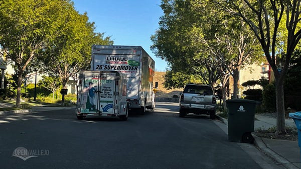 A photograph of a U-Haul truck and trailer driving down a tree-lined residential street.