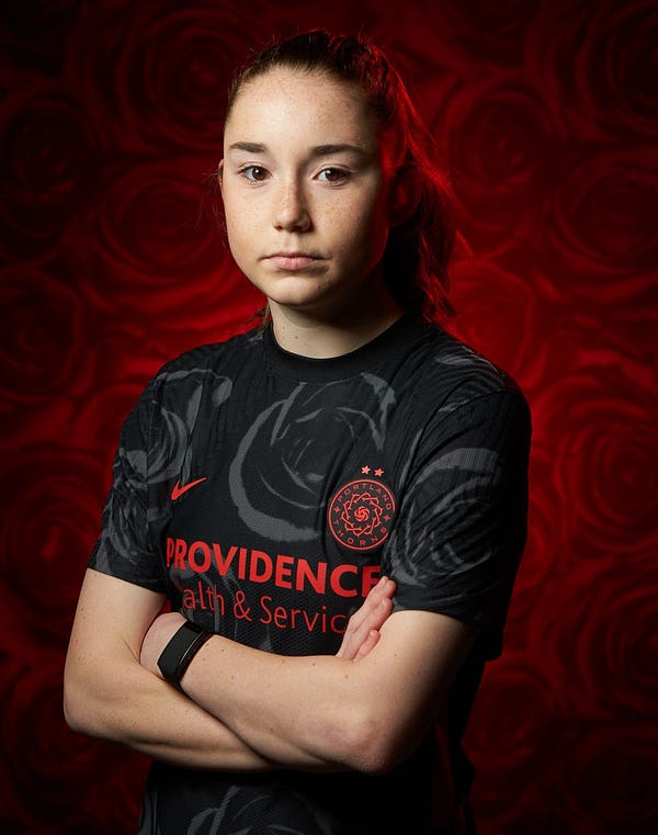 Olivia Moultrie poses with her arms crossed in a black Thorns kit with a background of red roses behind her