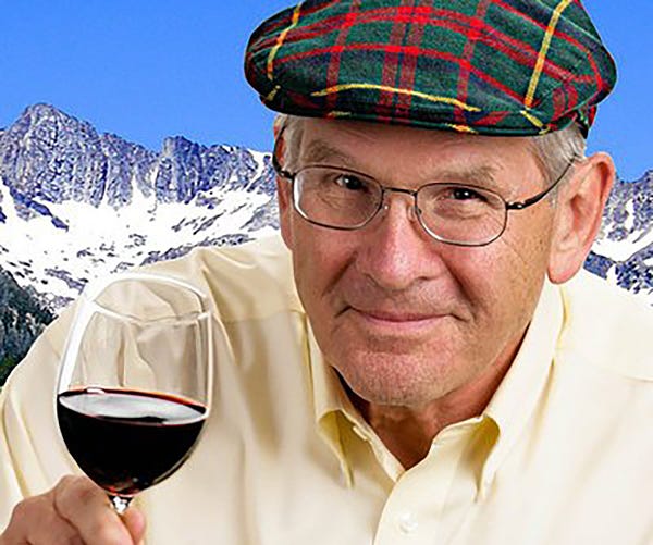 Rip McIntosh is seen wearing a plaid golf cap, glasses, and a yellow shirt. He's holding up a glass of red wine. Snow-covered mountains appear behind him in the background.