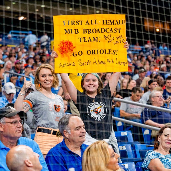 Two fans at the game holding up a sign that says "First all female MLB broadcast team! But not the last. Go Orioles!