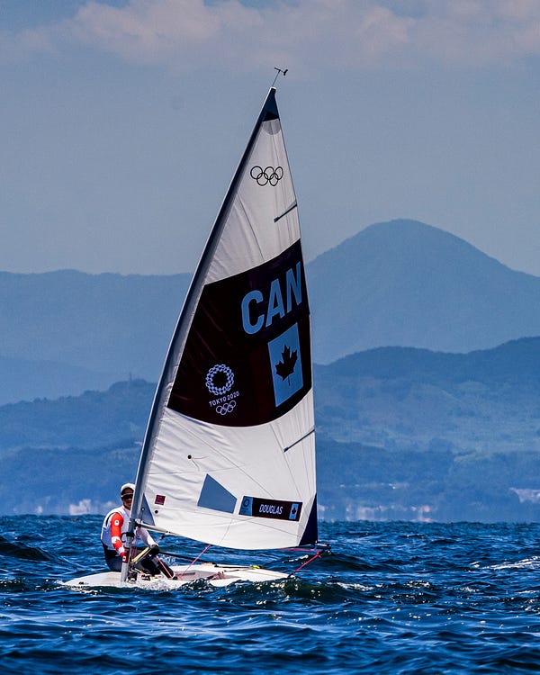 A wide shot of Sarah Douglas on her boat in the water during the race. Behind her, are mountains.