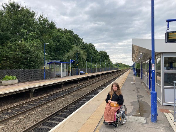 Katie, a white woman with long light-brown hair, is sat in her pink manual wheelchair on a platform at an outdoor train station. The platform is empty with no one else about. The railway track goes off behind her into the distance.