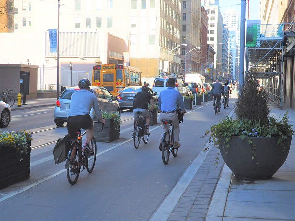 People cycle westbound on Richmond Street in a cycle track separated from vehicular traffic by planters and bollards. 