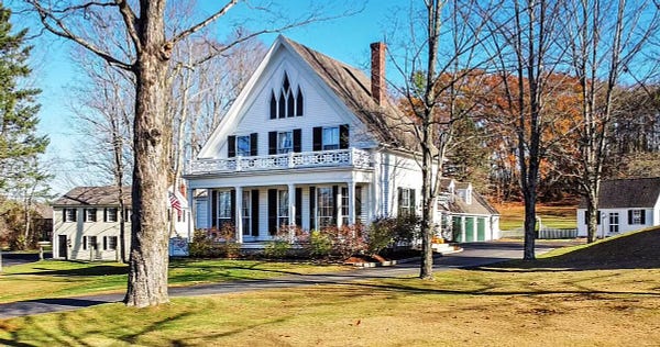 A white New England house with black shutters and a fancy central window, looking to be mid-19th century, with other buildings of similar age surrounding.