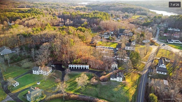 Aerial view of New England village nestled among trees in the hills — white buildings with black shutters, sunlight on the hills.