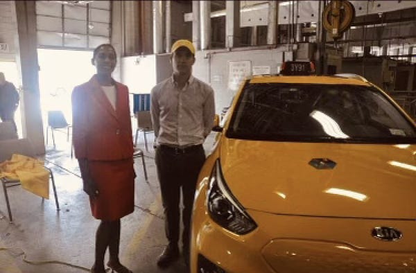 A woman wearing a red dress-suit smiles and stands alongside a man wearing a yellow hat in an inspection garage. To the right is  a Kia Niro EV taxi. 