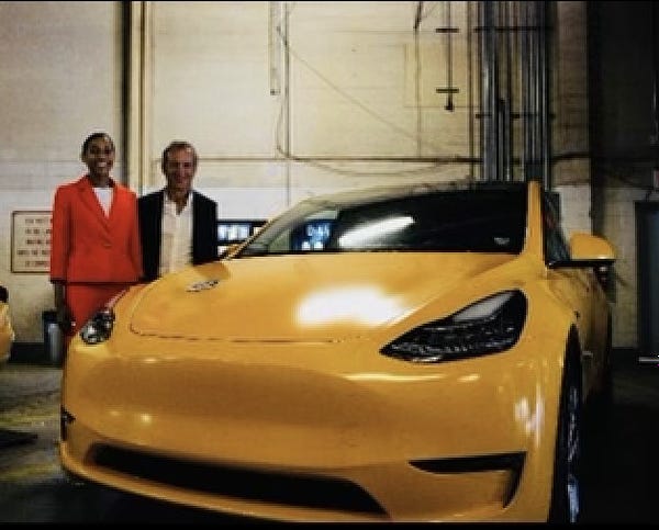 A woman wearing a red suit-dress is next to a man wearing an open blazer, to the left of a Tesla Model Y. There is a medallion tin on the front.
