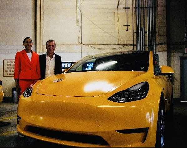 A woman wearing a red suit dress and a man wearing an open blazer stand to the left of a Tesla Model Y in a garage.