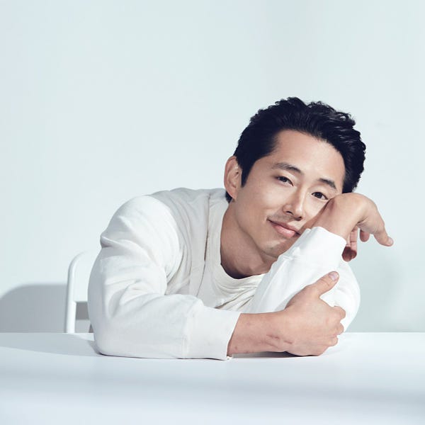 Steven Yeun in all white leaning on a white table. He is sitting in front of a white background.