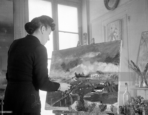 black and white photography of Poulain painting. she is on the left and we can see her back. she wears tight black suit (?) and has hair in tight bun. she faces a painted canvas (on the right) with a brush in her right hand. there is a getty images watermark.