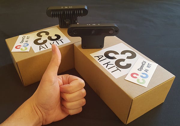 Two OpenCV OAD-D devices and their packages near a thumbs up.