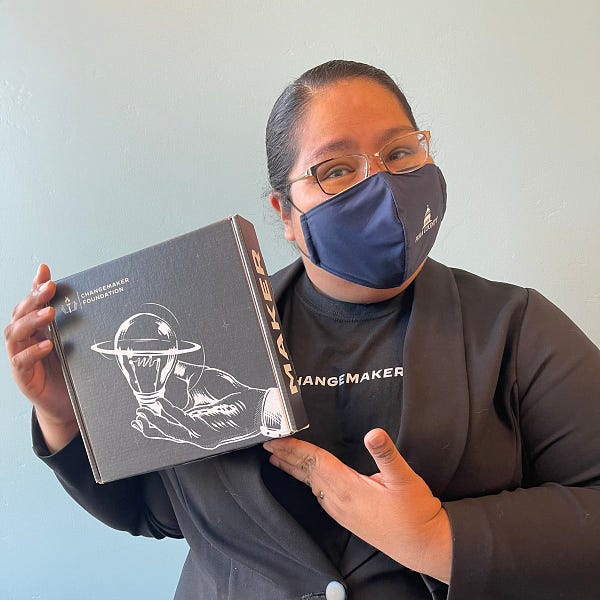Image description: Gabriella is holding up a small square box that has a pencil drawing on it of a hand holding a lightbulb. The lightbulb has a ring around it evoking the planet Saturn or movement. The box has a logo that reads “Changemaker Foundation. Gabriella is wearing a black shirt that says, “Changemaker” in white, a jacket, glasses and a blue face mask with the Pima County logo. She has her hair tied back into a bun.