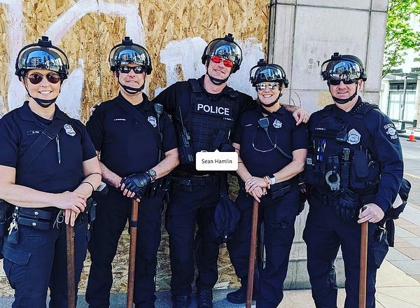 Image ID. Five Seattle police (three men and two women) wearing dark blue uniforms, sunglasses and riot helmets pose for a group photo downtown. Four of them have their arms folded and are leaning on long wooden clubs. The officer in the middle has his arms around the two to either side of him. There is a Facebook tag just below his chest that reads "Sean Hamlin"