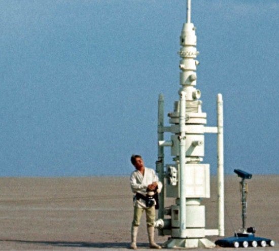 A scene from Star Wars where a person stands in the desert next to a tall round device.