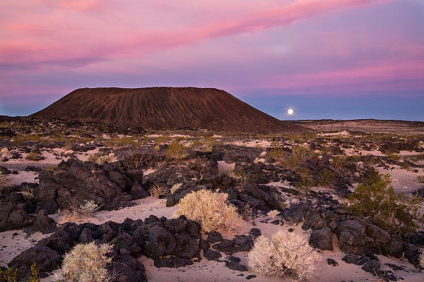 A distant volcanic mesa rises up from the rocky desert. A moon is just on the horizon of the sky. 