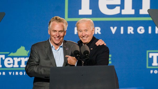 Terry McAuliffe (left) and President  Joe Biden (right) hug from the side behind a podium. President Biden is pointing his left hand at Terry's chest. Both are smiling. The blue backdrop behind them reads "TERRY FOR VIRGINIA."