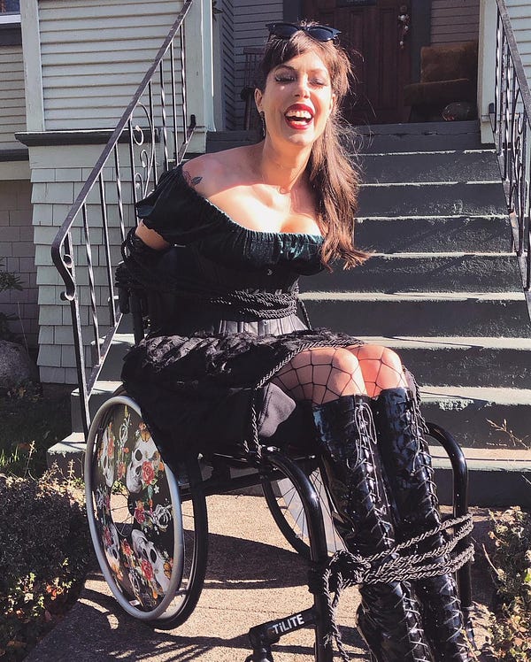 ID: Tara is a disabled woman with CRPS, using her rockin’ wheelchair. For Halloween she is faux bound to her wheelchair with rope. Here she is laughing.