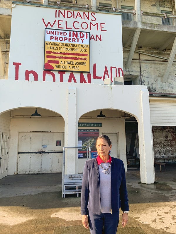Secretary Haaland stands in front of a sign with Indians Welcome graffiti on Alcatraz Island.