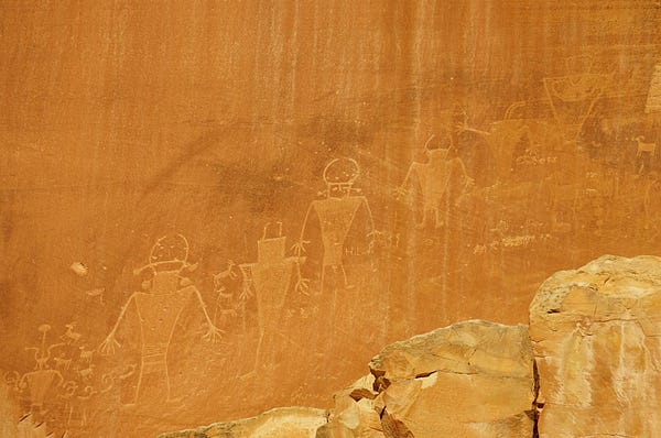 Human-like images and bighorn sheep images carved into a flat, vertical wall of orange sandstone.