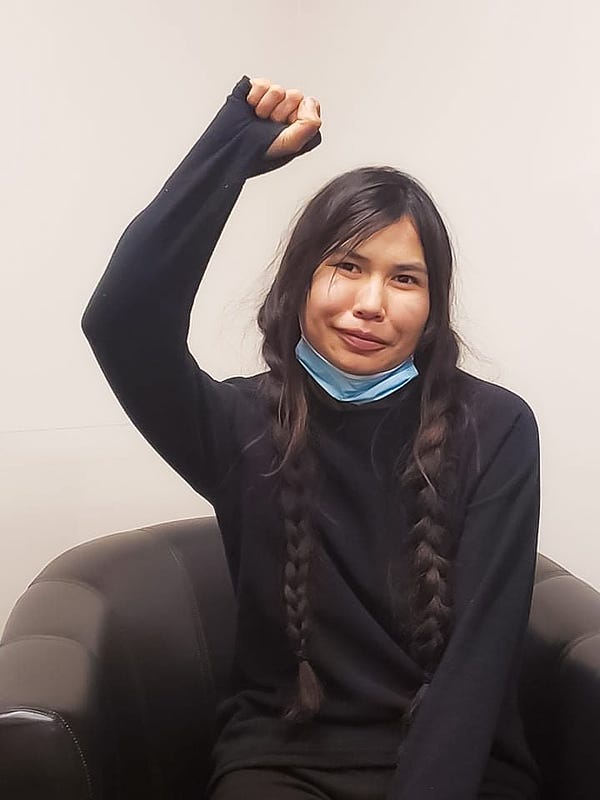 Image of Dinï ze’ Woos’ daughter Jocelyn Alec on a black armchair wearing black hoodie, blue mask pulled down, two braids, and right fist raised.