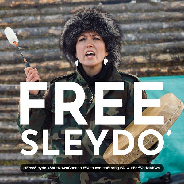 Picture of Sleydo' in front at Coyote Camp while singing and drumming, with drum in left hand. Text reads: Free Sleydo' in large white font and with hashtags #FreeSleydo #ShutDownCanada #WetsuwetenStrong underneath