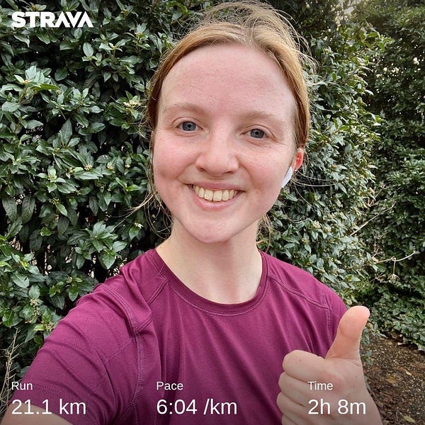 Charlie is wearing a burgundy t shirt and is smiling with her thumbs up. She has dark blonde hair and is wearing AirPods. At the bottom, there is white writing with the distance - 21.1km - the pace - 6.04 per km and time (2 hours 8 minutes). There’s a green bush behind her.