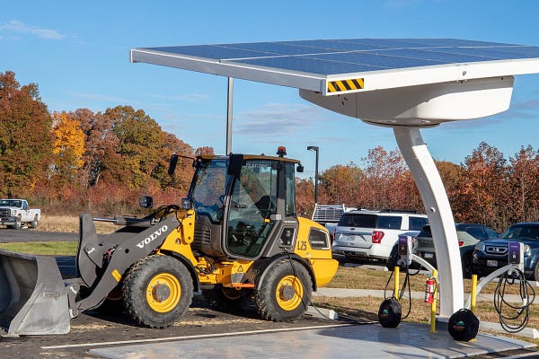 Electric equipment and solar-powered electric vehicle charging system in a parking lot with autumnal vegetation in the background and blue sky and white clouds above.