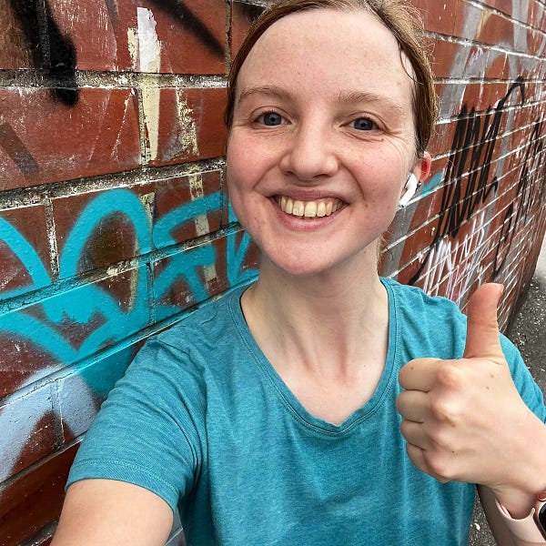 Charlie is wearing a sky blue T shirt and has her thumb up. She is smiling. There is a brown wall with graffiti on behind her.