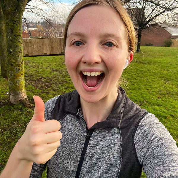 Charlie - a 28 year old woman with dark blonde hair and blue eyes - is wearing a grey jacket. She has her mouth open excitedly and has her thumb up. Behind her is a green field with trees.