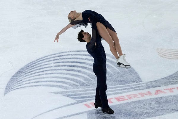 Two figure skaters perform on the ice. The male figure skater is lifting the woman over his head