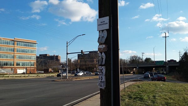 Two sets of ghost shoes and plaques on a utility pole next to an off-ramp, near the Beltway overpass over New Hampshire Ave. The top plaque says Mark Henderson.