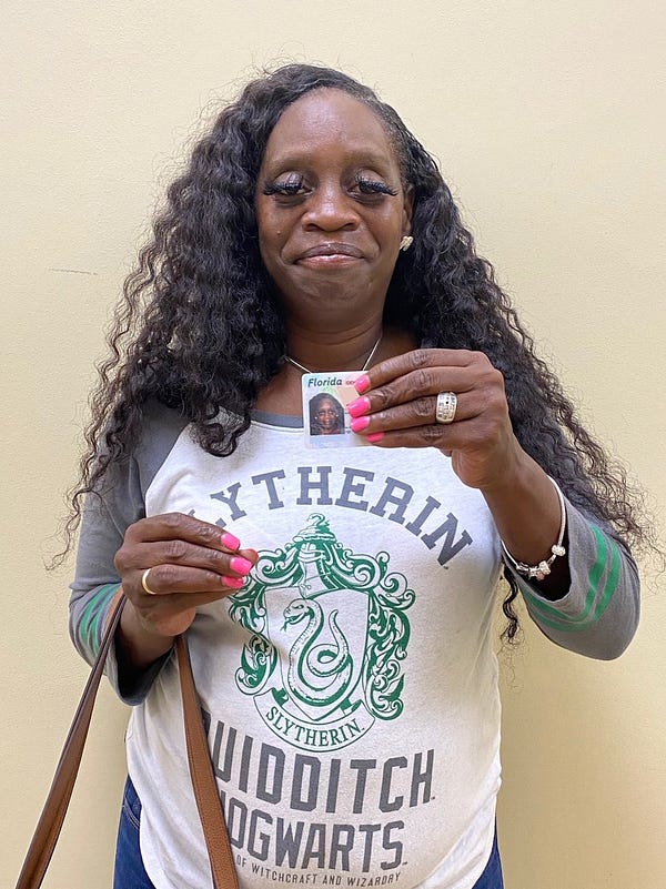A Black woman with long, wavy hair and bright pink nails displays her Florida photo ID. She is wearing a Harry Potter shirt and several pieces of gold jewelry.