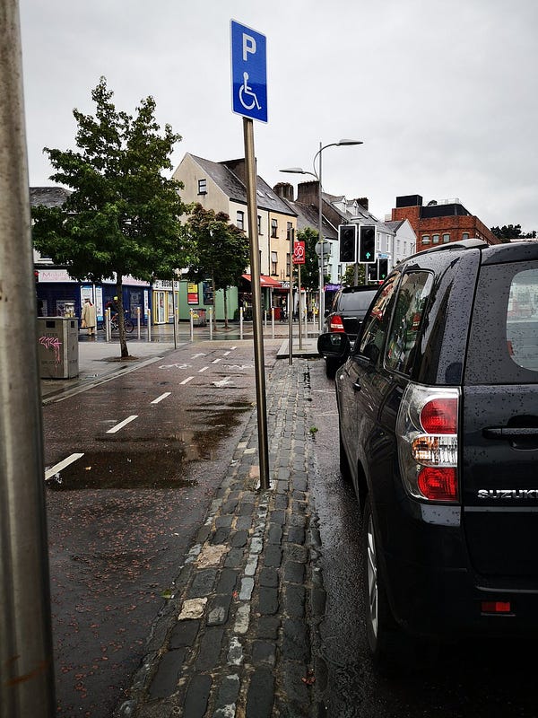 An on-street disability parking bay moved away from the path to accommodate a contra-flow cycle lane. Disability signage pole is placed on passenger side of vehicle erected on a cobbled buffer zone with a gradient.