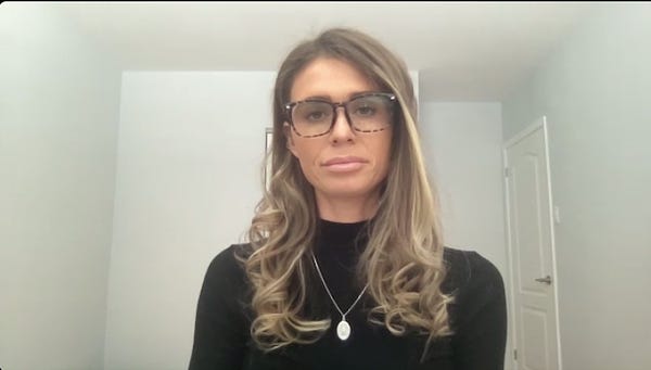 A Zoom window-type window showing Faith Goldy in a black sweater with a silver pendant and large glasses, sitting in a blank white room. She looks impassive.