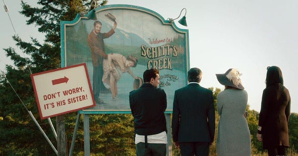 Schitt’s Creek town sign with a sign reading “Don’t worry, it’s his sister!” with an arrow pointing to a man standing behind a woman bending over.