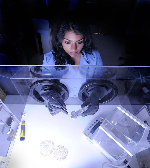 Woman of color working a lab with gloves. She is wearing a blue shirt and has her dark hair down. 
