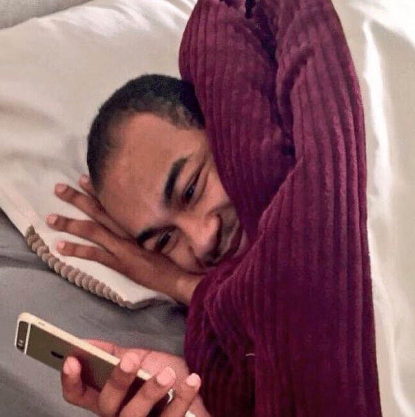 Meme of a man laying in bed under the covers and smiling at his phone.